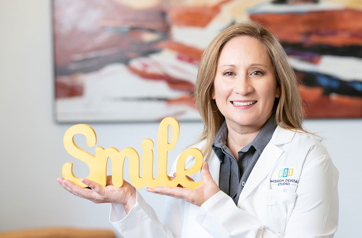 Dr. Shiosaky Promoting Healthy Smiles – Mission Dental Studio  Dr. Shiosaky, a smiling female dentist in a white coat, holds a yellow "Smile" sign while standing in front of colorful abstract artwork at Mission Dental Studio